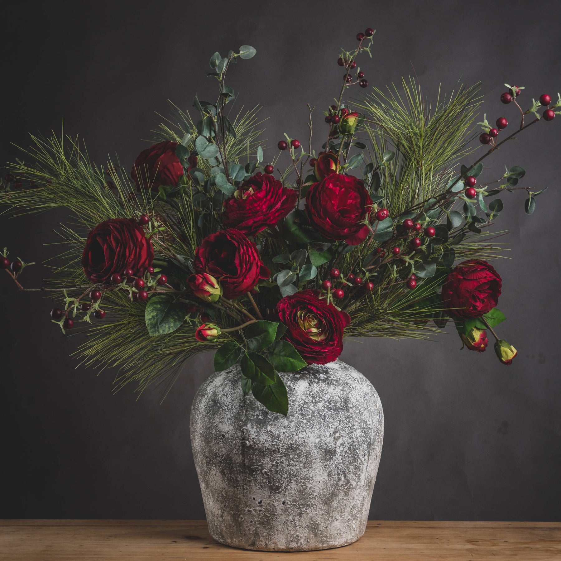 Stone-grey ceramic urn vase with deep red floral arrangement against dark backdrop.