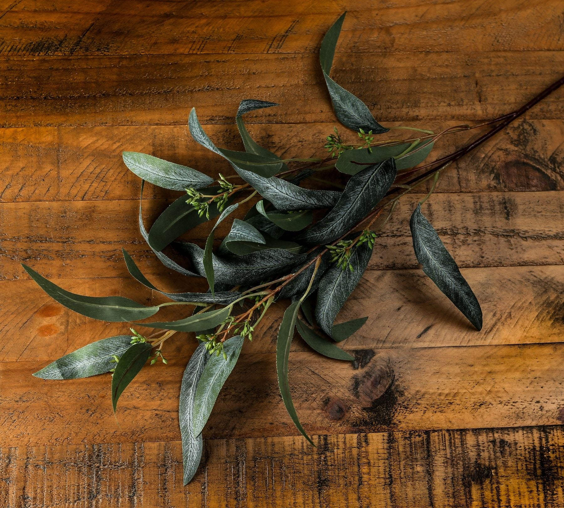 Overhead view of Seeded Eucalyptus Faux Spray stems laid on a reclaimed wooden table, highlighting olive-green leaves and muted seed bud accents.