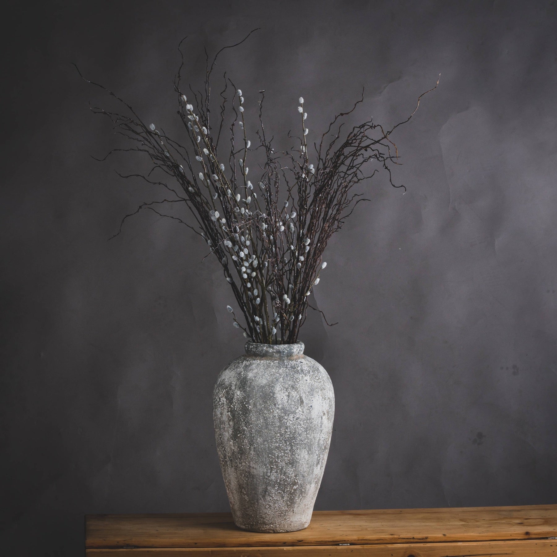 Grey stoneware vase styled with white faux willow stems, shown on a neutral surface. Sculptural form and tonal texture highlight calm, minimalist decor.