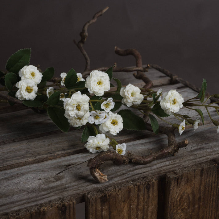 Single stem of White Wild Meadow Rose Faux Spray laid horizontally on weathered timber, highlighting realistic petal texture.